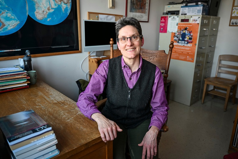 Claire Wendland sits, smiling, in wooden chair in an office with a worn wooden desk and maps and art on the walls. Photo by Althea Dotzour  / UW–Madison